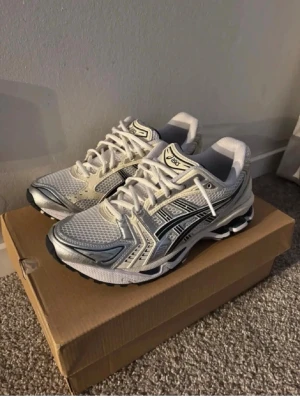 Vita ASICS sneakers med silvriga detaljer - Snygga vita ASICS sneakers med silvriga och svarta detaljer. Ovandelen är i mesh för bra andningsförmåga och har klassiska ASICS-loggor på sidorna. Sulan är robust och ger bra stöd, perfekt för en sportig och trendig look.