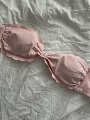 Ljusrosa bandeau bikiniöverdel - Söt ljusrosa bikiniöverdel i bandeau-modell med vågiga kanter. Bikinin har en knut i mitten och avtagbara axelband. Perfekt för dig som gillar en feminin och trendig stil på stranden.