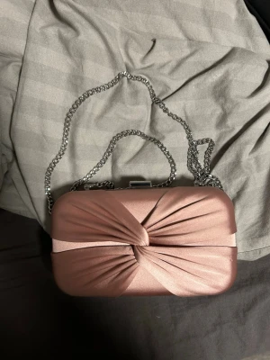 Rosa kuvertväska med silverkedja från anna field -  kuvertväska i ljusrosa satin med dekorativ twistad front och silverfärgad kedja. Väskan har hårt skal och metallspänne upptill. (Kedjan går att ta av)