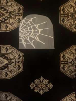 Grå mössa med spindelnätsmönster - Snygg grå mössa med ett stort vitt spindelnätsmönster på ena sidan. Mössan har en klassisk, rundad form och är tillverkad i ett mjukt och stretchigt material som passar perfekt för kyliga dagar. Perfekt accessoar för dig som gillar unika detaljer.