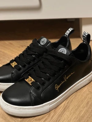 Svarta Versace sneakers med gulddetaljer - Snygga svarta sneakers från Versace i läder med vita sulor och guldfärgade detaljer. Skorna har svart snörning, Versace-logga på tungan och hälen samt Gianni Versace-signatur i guld på sidan. Grekiskt mönster på hälen för extra stilpoäng. Helt nya, aldrig använda. 