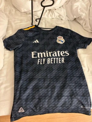 Real Madrid bortatröja Adidas - Mörkblå Real Madrid bortatröja från Adidas med gul detalj på axeln och klubbmärke på bröstet. Tröjan har korta ärmar, rund hals och tryck med 'Emirates Fly Better' på framsidan. Tillverkad i lätt och ventilerande material.