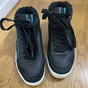 Svarta Puma sneakers med blå detaljer - Säljer ett par svarta Puma sneakers med blå detaljer och vit sula. Skorna har snörning, rund tå och är fodrade med mjukt material för extra värme. Perfekta för dig som vill ha en sportig look med lite extra komfort under kallare dagar.