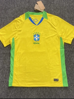 Brasilien fotbollströja gul/grön - Snygg gul fotbollströja från Brasilien med gröna detaljer på sidorna och runt kragen. Tröjan har CBF-loggan och fem stjärnor på bröstet. Tillverkad i lätt och ventilerande syntetmaterial, perfekt för träning eller match.