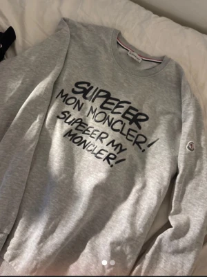 Grå sweatshirt från Moncler - Snygg grå sweatshirt från Moncler med tryck i svart text på framsidan där det står 'SUPEEER MON MONCLER!'. Tröjan har rund halsringning, långa ärmar och en liten Moncler-logga på ärmen. Perfekt för en avslappnad och trendig look.