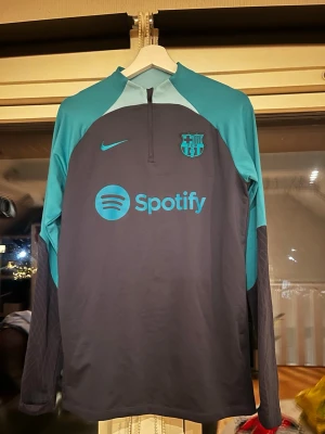Nike Barcelona träningströja blå/svart - Snygg långärmad träningströja från Nike med FC Barcelona-logga och Spotify sponsortryck. Tröjan är svart med turkosa detaljer och har halv dragkedja framtill. Tillverkad i Dri-FIT material som andas, perfekt för träning eller uppvärmning.