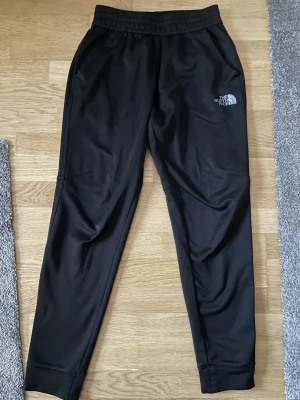 Svarta mjukisbyxor från The North Face - Svarta mjukisbyxor från The North Face med elastisk midja och smal passform. Byxorna har två sidofickor och logga tryckt på både fram- och baksida. Tillverkade i mjukt och stretchigt material som är perfekt för chill eller träning.