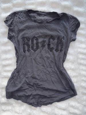 Grå t-shirt med ROCK-tryck från Zadig & Voltaire - Cool grå t-shirt från Zadig & Voltaire med glittrigt ROCK-tryck och blixtdetalj framtill som ej säljs längre. T-shirten har rund halsringning och korta ärmar. Perfekt för dig som gillar en edgy och avslappnad stil. Köpt second hand och något nopprig. Storlek XS-S