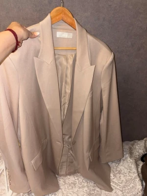 Beige kavaj från Ellos och rutig kavaj - Två snygga kavajer: en beige från Ellos med klassisk passform och slät yta, samt en brunrutig kavaj med svart foder och dubbelknäppning. Båda har fickor fram och markerade slag. Perfekta för att lyfta din outfit med en stilren touch.