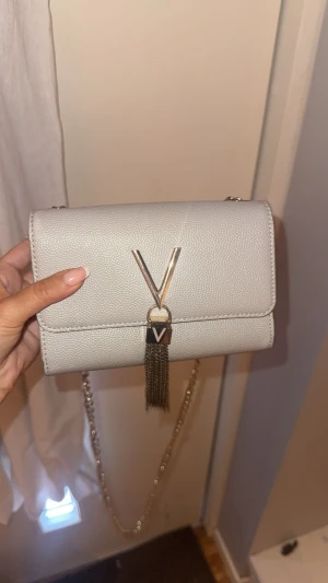 Vit axelväska från Valentino - Snygg vit axelväska från Valentino mario med guldfärgad kedjerem och dekorativt V-spänne framtill. Väskan har en elegant tofsdetalj och är tillverkad i ett strukturerat konstläder. Perfekt storlek för det viktigaste och ger en lyxig touch till din outfit.