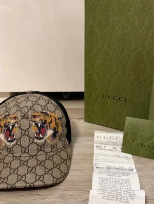 Gucci keps med tigerbrodyr - Säljer en snygg Gucci keps med klassiskt GG-mönster i beige och brunt, prydd med två broderade tigrar framtill. Justerbar rem med grön-röd rand bak och svart mesh på insidan för extra komfort. Perfekt för dig som vill sticka ut med lyxig streetstyle.