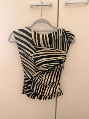 Zebramönstrad ärmlös topp - Snygg ärmlös topp med zebramönster i svart och beige. Toppen har en figurnära passform och coola diagonala ränder som ger en unik look. Perfekt att styla med jeans eller kjol för en trendig outfit.