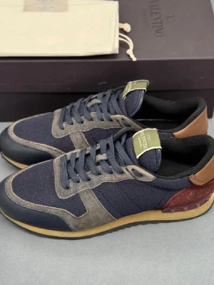 Valentino Garavani blå sneakers - Snygga sneakers från Valentino Garavani i marinblå mesh med grå mockadetaljer och mörkblå läderpartier. Skorna har bruna och vinröda detaljer vid hälen samt gummisula. Klassisk låg modell med märkets logga på plösen. Inga defekter alls bara lite smuts där fram som jag tvättar bort vid köp