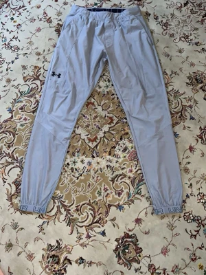 Ljusgrå mjukisbyxor från Under Armour - Säljer ett par ljusgrå mjukisbyxor från Under Armour med smal passform och resår i bensluten där det står 'UNDER ARMOUR'. Byxorna har en diskret logga på vänster lår och är gjorda i ett lätt och stretchigt material som känns riktigt skönt.