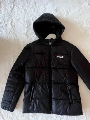Svart pufferjacka från FILA med huva - Säljer en ny svart pufferjacka från FILA med huva och dragkedja framtill. Jackan har två fickor med dragkedja