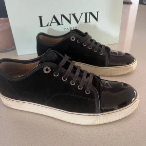 Svarta sneakers från Lanvin med lackdetalj - Snygga svarta sneakers från Lanvin med ovandel i mocka och glansig lackad tå. Skorna har vita gummisulor och klassisk snörning. Perfekt för dig som gillar stilrena men ändå unika detaljer. Kommer med originalkartong och dustbag.