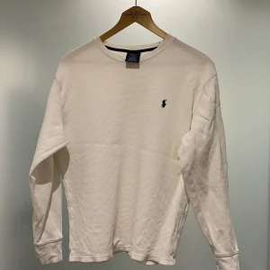 Vit långärmad tröja Polo Ralph Lauren - Vit långärmad tröja från Polo Ralph Lauren i 100% bomull. Tröjan har en diskret struktur, rund halsringning och den klassiska broderade loggan på bröstet. Enkel och stilren modell som passar perfekt till jeans. 
