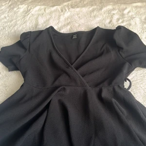 Svart omlotttöja från SHEIN - Snygg svart omlottklänning från SHEIN i storlek M. Klänningen har korta puffärmar, v-ringad hals och knyt i sidan för en feminin siluett. Materialet är mjukt och lätt, perfekt för dig som gillar stilrena och enkla plagg med en twist.