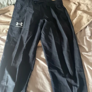 Svarta träningsbyxor Under Armour YXL - Svarta träningsbyxor från Under Armour i storlek YXL. Byxorna har elastisk midja, dragkedjor vid bensluten och logga på ena benet. Lätta och luftiga, perfekta för sport eller chill. Loose fit för skön rörelsefrihet.
