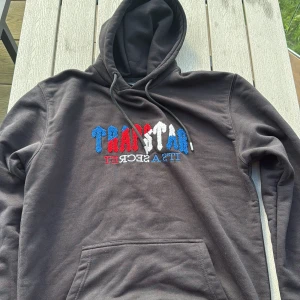 Svart Trapstar hoodie med brodyr - Svart hoodie från Trapstar med broderad logga i blått, rött och vitt på bröstet. Klassisk känguruficka och justerbar huva med snören. Mjuk bomullskänsla, perfekt för en chill streetwear-look.