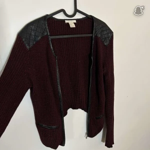 Vinröd cardigan jacka från H&M - Snygg vinröd stickad cardigan jacka från H&M med svarta quiltade axelpartier i skinnimitation och dragkedja framtill. Jackan har två fickor med svarta detaljer och långärmad passform. Perfekt för lager-på-lager-stil under höst och vinter.