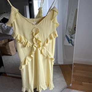 Gul volangklänning från H&M XS - Söt ljusgul klänning från H&M i storlek XS med volangdetaljer längs axlar, fram och nederkant. Klänningen har smala axelband och är tillverkad i ett lätt, strukturerat tyg som känns som bomull. Perfekt för sommarens alla tillfällen!