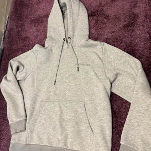 Grå hoodie från Peak Performance - Snygg grå hoodie från Peak Performance med broderad logga på bröstet. Klassisk känguruficka, huva med snörning och ribbade muddar. Perfekt för chill dagar eller när du vill ha en enkel och stilren look. Är medium men sitter mer som small