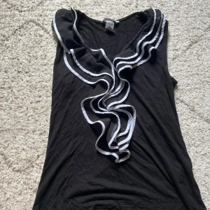 Svart linnetop med volang  från H&M XS - Svart ärmlös topp från H&M i storlek XS med snygga volangdetaljer framtill som har vita kanter. Toppen har en v-ringad hals och är tillverkad i ett mjukt, lätt material. Har fått den av en kompis men inte andvänt så mycket själv.