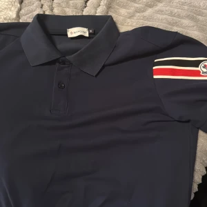Mörkblå Moncler t-shirt med ränder - Snygg mörkblå t-shirt från Moncler med klassisk krage och två knappar framtill. På ena ärmen finns broderad Moncler-logga och coola ränder i vitt, svart och rött. Perfekt för dig som gillar stilrena märkesplagg med lite extra detaljer.