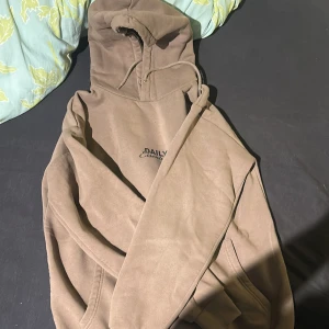 Beige hoodie från Daily Essentials - Snygg beige hoodie från Daily Essentials med stor svart logga och text på ryggen. Tröjan har huva med snörning och känguruficka framtill. Perfekt för en chill och avslappnad stil. Mjuk och skön, tillverkad i bomullsmix.