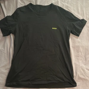 Svart t-shirt från Hugo Boss i bomull - Svart t-shirt från BOSS med diskret logga i gult på bröstet. Klassisk rund halsringning och korta ärmar. Tillverkad i mjuk bomull som känns skön mot huden. Perfekt för en clean och stilren look. Pris kan diskuteras.
