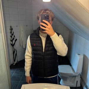 Svart dunväst från Polo Ralph Lauren - Svart dunväst från Polo Ralph Lauren med horisontella sömmar och röd broderad logga på bröstet. Västen har dragkedja framtill och två fickor med dragkedja. Perfekt för lager-på-lager och snygg till hoodie eller stickad tröja.