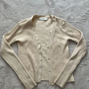 Beige ribbad kofta från SAMSA Collection - Snygg beige ribbad kofta från SAMSA Collection i storlek S. Koftan har lång ärm, v-ringning och knappar framtill. Perfekt att slänga över en topp för en chill och stilren look. Materialet känns mjukt och stretchigt.