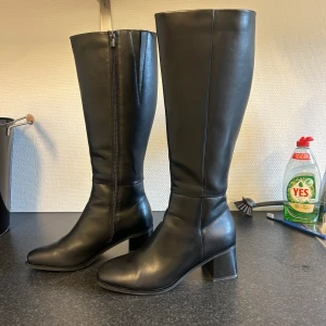 Svarta höga stövlar i skinn - Snygga svarta stövlar i  skinn med dragkedja på insidan och låg klack. Klassisk design med rund tå och slät yta. 