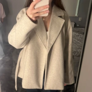 Beige kappa med bälte från vero Moda  - Säljer en snygg beige kappa från vero Moda med bred krage och bälte i midjan. Jackan har en loose passform och är tillverkad i ett mjukt ullblandat material. Perfekt för höst och vår när du vill ha en stilren look.