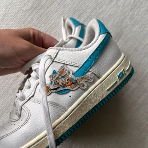 Nike Air Force 1 Space Jam sneakers - Unika Nike Air Force 1 sneakers i vitt skinn med turkosa detaljer och Space Jam-motiv på sidorna. Skorna har klassisk snörning, rund tå och platt sula i turkos. Perfekta för dig som vill sticka ut med en cool och lekfull stil.