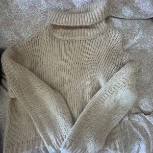 Beige stickad polotröja med detaljer - Mysig beige stickad polotröja med grovstickad känsla och lång ärm. Tröjan har en hög polokrage och snygga guldfärgade tråddetaljer längs ärmsluten som ger en extra touch. Perfekt för kalla dagar och enkel att styla till jeans eller kjol.