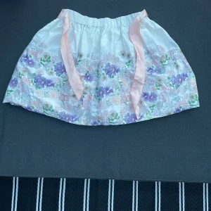 Vit blommig kortkjol med blommor  - Supersöt vit kortkjol med blommigt mönster i lila, rosa och grönt. Kjolen har en ljusrosa rosett och band i midjan som ger en gullig touch. Perfekt för vår och sommar när du vill ha en romantisk och lekfull stil.