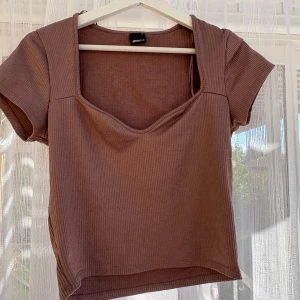 Gammelrosa ribbad topp från Gina Tricot - Säljer en gammelrosa ribbad topp från Gina Tricot med korta ärmar och snygg v-ringning. Toppen har en croppad passform och är tillverkad i mjuk bomullsmix. Perfekt att styla med jeans eller kjol för en trendig look.