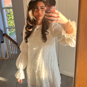 Vit kort klänning med volangärmar från Gina  - Superfin vit kort klänning med volangärmar och diskret blommönster i tyget. Klänningen har en lös passform och är tillverkad i ett lätt material som ger en romantisk och luftig känsla. Perfekt för student eller sommarens alla tillfällen!