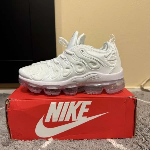 Nike Air Vapormax Plus vita sneakers - Nike Air Vapormax Plus i helvitt med futuristisk design och genomskinlig sula. Ovandel i syntetmaterial med vågiga detaljer och snörning. Sulan har synliga luftbubblor för maxad komfort och cool look. Perfekta för dig som vill sticka ut med stilrena sneakers.