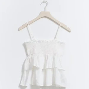 Vit volangtop från Gina Tricot - Supersöt vit topp från Gina Tricot med smala justerbara axelband ( avtagsbara) och volanger. Smockad upptill och luftig i bomull. Enkel att matcha med jeans och en kofta! Antigen som den är eller en bandeu topp! Skriv om du är intresserad eller har några frågor!🫶🍁