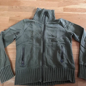 Olivgrön kofta från Gina Tricot - Olivgrön kofta från Gina Tricot i 100% bomull. Tröjan har ribbade muddar och hög krage, hel dragkedja framtill och två fickor med dragkedja.