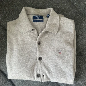 Grå kofta i lammull från Gant - Grå kofta från Gant i superfin lammull. Klassisk design med krage, knappar hela vägen och broderad logga på bröstet. Perfekt för lager-på-lager och funkar året om. Tröjan är i bra skick men har ett litet hål på armen plus några hål på ryggen. Inget man lägger jätte stor bemärkelse till.