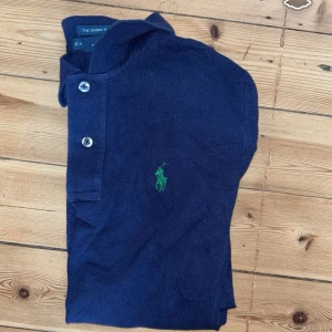 Marinblå pikétröja Ralph Lauren XS - Snygg marinblå pikétröja från Ralph Lauren i modellen The Skinny Polo. Tröjan har korta ärmar, klassisk krage och knäppning med två knappar framtill. Ikoniska gröna Polo-loggan broderad på bröstet. Tillverkad i mjuk bomull för en bekväm känsla.