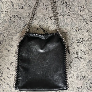 TIAMO VÄSKA - Cool svart axelväska från Tiamo i skinnimitation med silverfärgade kedjor längs kanterna och som axelrem. Väskan har dragkedja baktill. Bra skick förut att några detaljer gått upp(bild 3)🤍