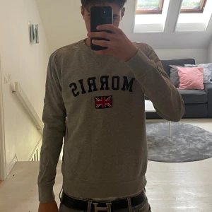 Grå sweatshirt från Morris - Säljer en grå sweatshirt från Morris med broderad logga och brittisk flagga på bröstet. Tröjan har rund hals, långa ärmar och ribbade muddar. Perfekt för en chill och stilren look.