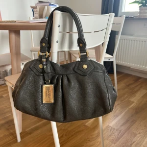 Grön handväska från Marc Jacobs - Snygg grön handväska från Marc by Marc Jacobs i äkta skinn med guldfärgade metalldetaljer och en stor logoplatta framtill. Väskan har två rejäla handtag, dragkedja upptill och ett coolt innerfoder med tryckt logga. Perfekt storlek för allt du behöver ha med dig.