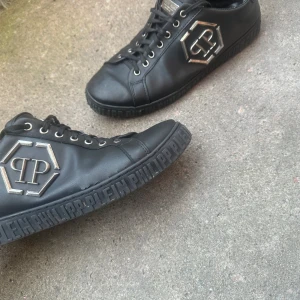 Svarta sneakers från Philipp Plein - Svarta sneakers i skinn från Philipp Plein med silverfärgad logga på sidan och präglad text runt sulan. Klassisk snörning och rund tå. Snygg och stilren modell med exklusiv känsla och coola metalldetaljer.