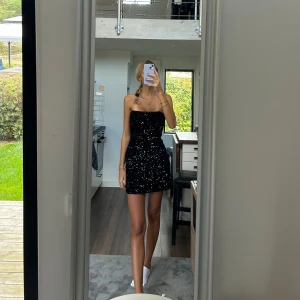 Svart paljettklänning axelbandslös - Axelbandslös svart kortklänning täckt av glittrande paljetter. Sitter tight och har en rak, enkel form som markerar midjan. Perfekt för att sticka ut med en riktigt festlig look eller som nyårsklänning.